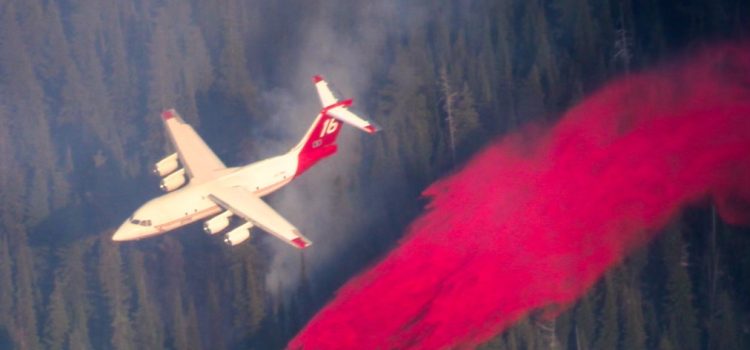 Een groot blusvliegtuig laat een brede rode sliert brandvertrager los boven een naaldbos waar de rook tussen de bomen hangt.